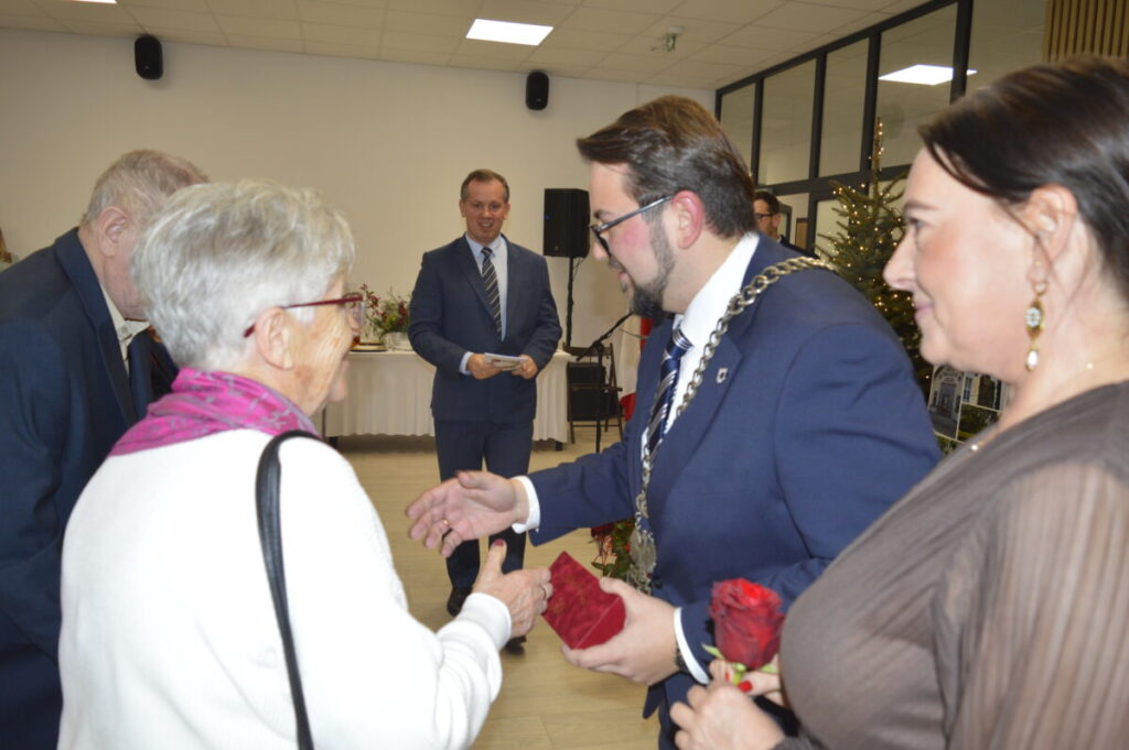 Spotkanie towarzyskie w jasnym pomieszczeniu. Mężczyzna w garniturze i łańcuchu wita starszą kobietę. W tle choinka i stół.