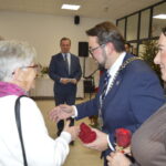 Spotkanie towarzyskie w jasnym pomieszczeniu. Mężczyzna w garniturze i łańcuchu wita starszą kobietę. W tle choinka i stół.