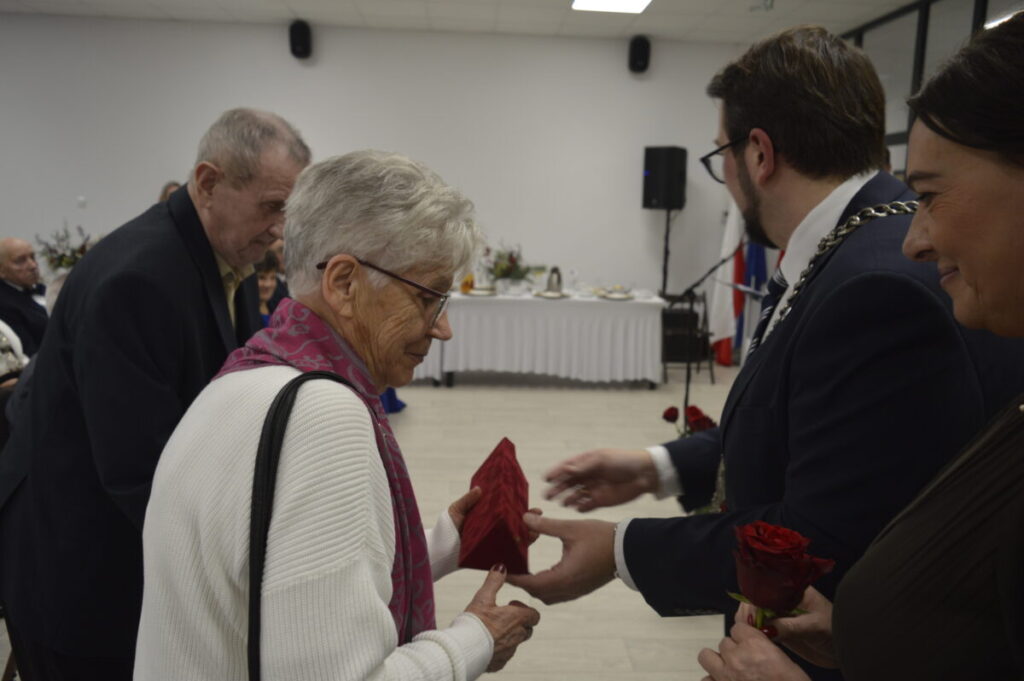 Grupa osób podczas uroczystości w sali. Starsza kobieta w białym swetrze otrzymuje prezent. W tle biały obrus i flagi.