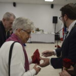 Grupa osób podczas uroczystości w sali. Starsza kobieta w białym swetrze otrzymuje prezent. W tle biały obrus i flagi.