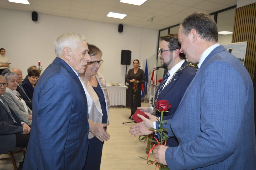 Starsza para w eleganckich ubraniach odbiera gratulacje i różę od mężczyzny w garniturze. W tle siedzą ludzie na krzesłach. Jasne wnętrze z dekoracjami.