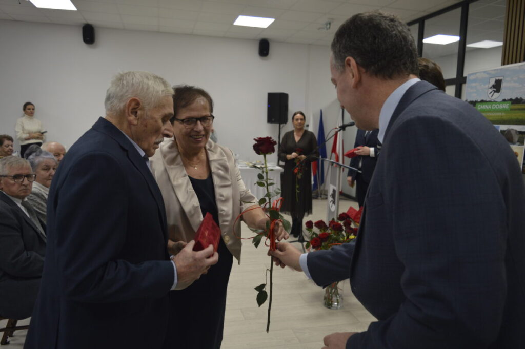 Grupa osób w formalnych strojach w jasnym pomieszczeniu. Mężczyzna wręcza czerwoną różę starszemu mężczyźnie i kobiecie. W tle siedzący ludzie i dekoracje.