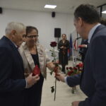 Grupa osób w formalnych strojach w jasnym pomieszczeniu. Mężczyzna wręcza czerwoną różę starszemu mężczyźnie i kobiecie. W tle siedzący ludzie i dekoracje.