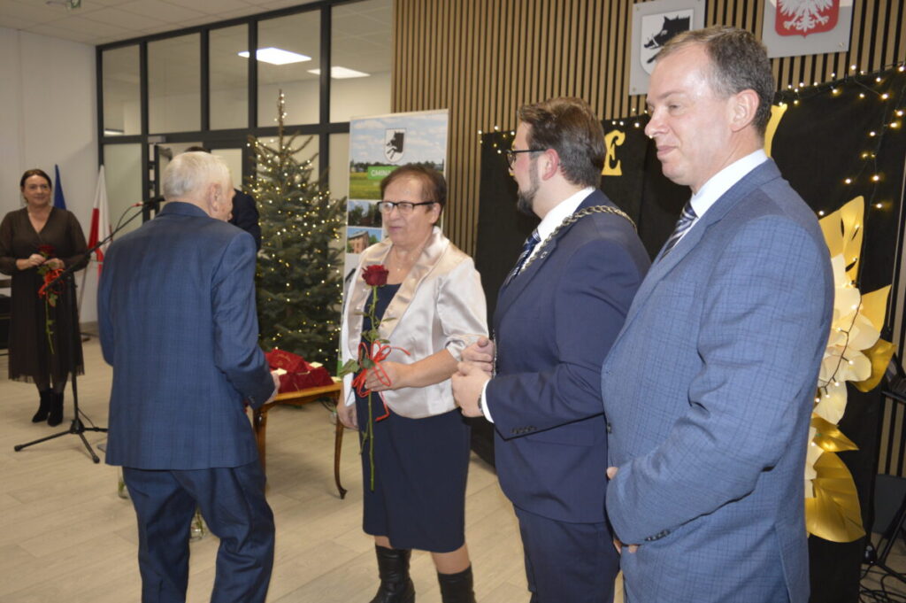 Grupa osób w eleganckich strojach stoi w jasnym pomieszczeniu z choinką. Za nimi czarne tło z żółtymi dekoracjami. Dwie osoby trzymają czerwone róże.