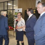 Grupa osób w eleganckich strojach stoi w jasnym pomieszczeniu z choinką. Za nimi czarne tło z żółtymi dekoracjami. Dwie osoby trzymają czerwone róże.