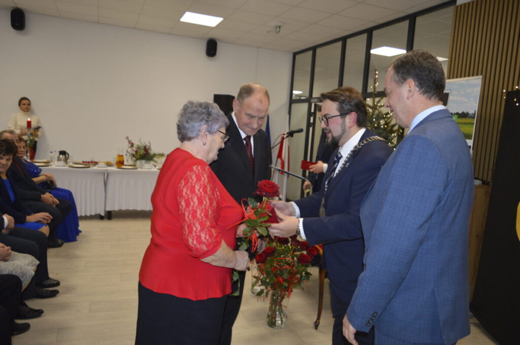 Grupa osób w eleganckich strojach, starsza kobieta w czerwieni otrzymuje kwiaty w jasnym pomieszczeniu. Tło: stół z białym obrusem, dekoracje, choinka. Klimat uroczysty, oficjalny.