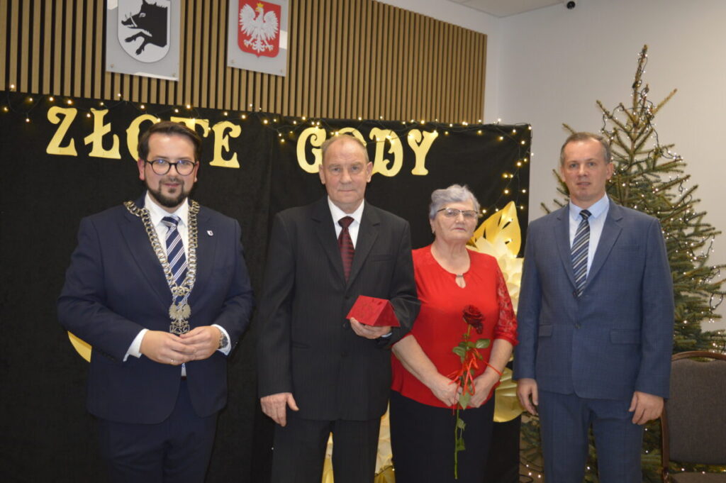 Na zdjęciu cztery osoby stojące przed czarnym tłem z napisem Złote Gody. Mężczyzna trzyma czerwoną kopertę, kobieta czerwoną różę. Obok nich choinka ozdobiona światełkami.