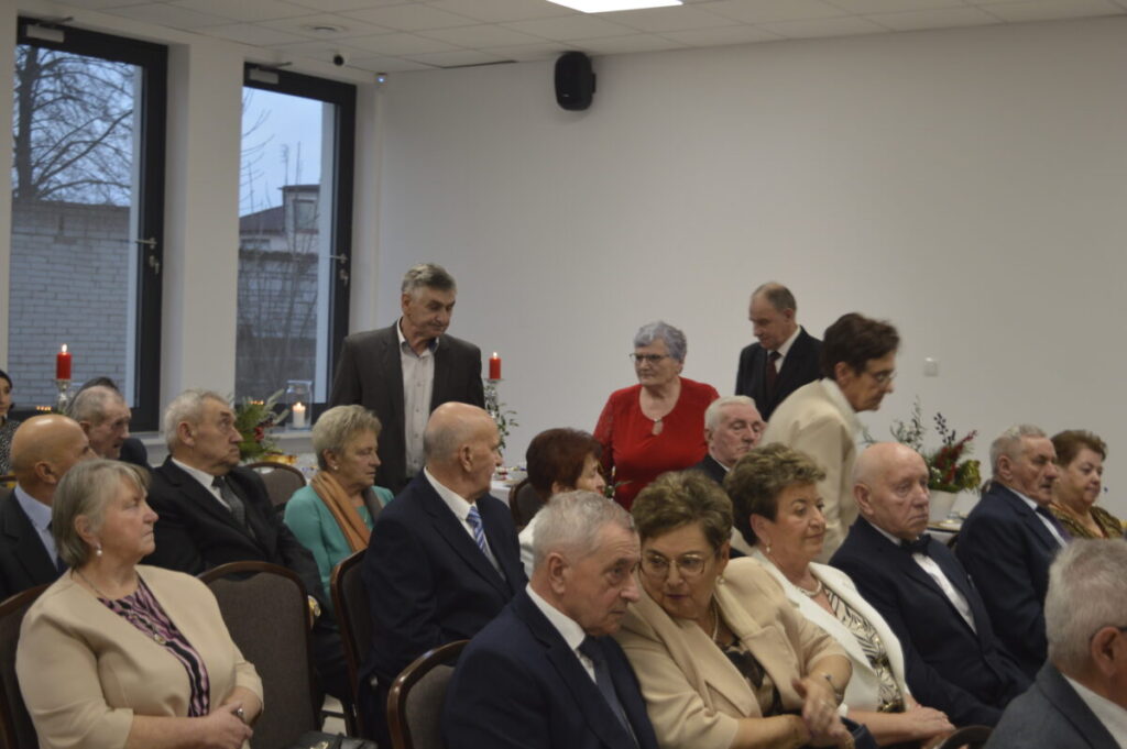 Grupa starszych osób siedzi na uroczystości w jadalni z dużymi oknami w tle. Wszyscy ubrani elegancko, atmosfera formalna. W tle widoczne są czerwone świece i dekoracje.