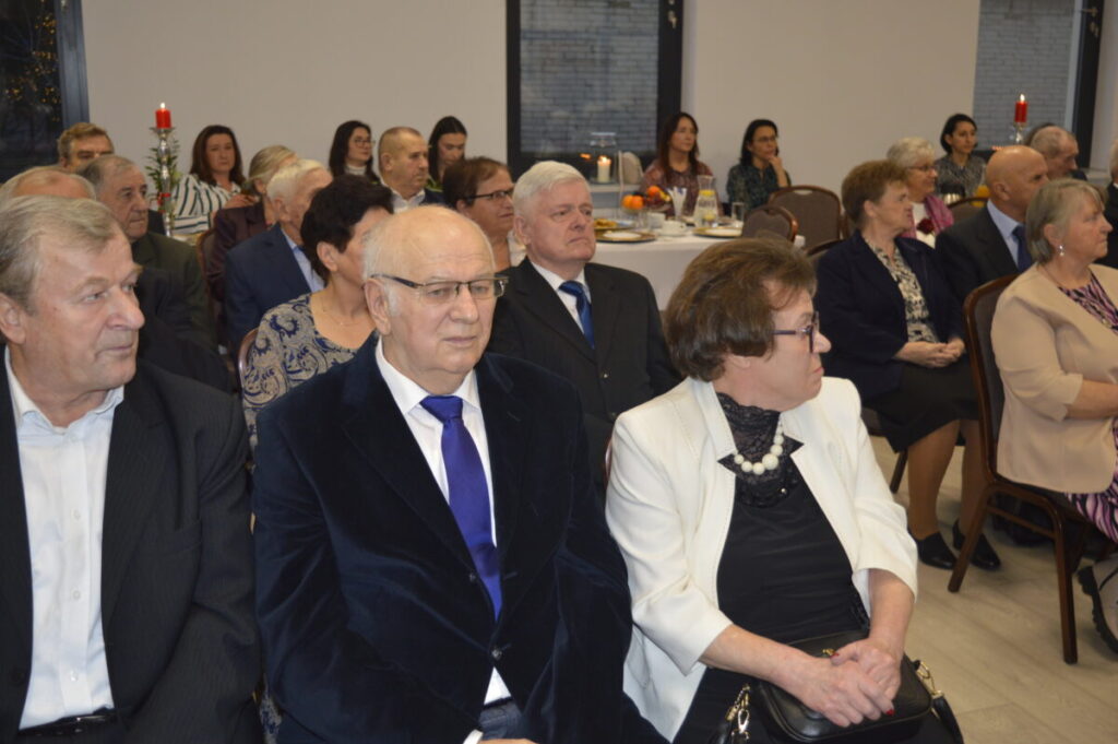 Grupa osób siedzi w eleganckiej sali, ubrani formalnie. Tło jasne z dekoracjami i stołem. Atmosfera oficjalna, uroczysta.