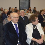 Grupa osób siedzi w eleganckiej sali, ubrani formalnie. Tło jasne z dekoracjami i stołem. Atmosfera oficjalna, uroczysta.