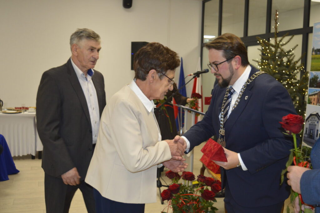 Na obrazie widoczne są trzy osoby w eleganckiej sali. Mężczyzna w garniturze wręcza starszej kobiecie prezent. W tle choinka i dekoracje świąteczne.