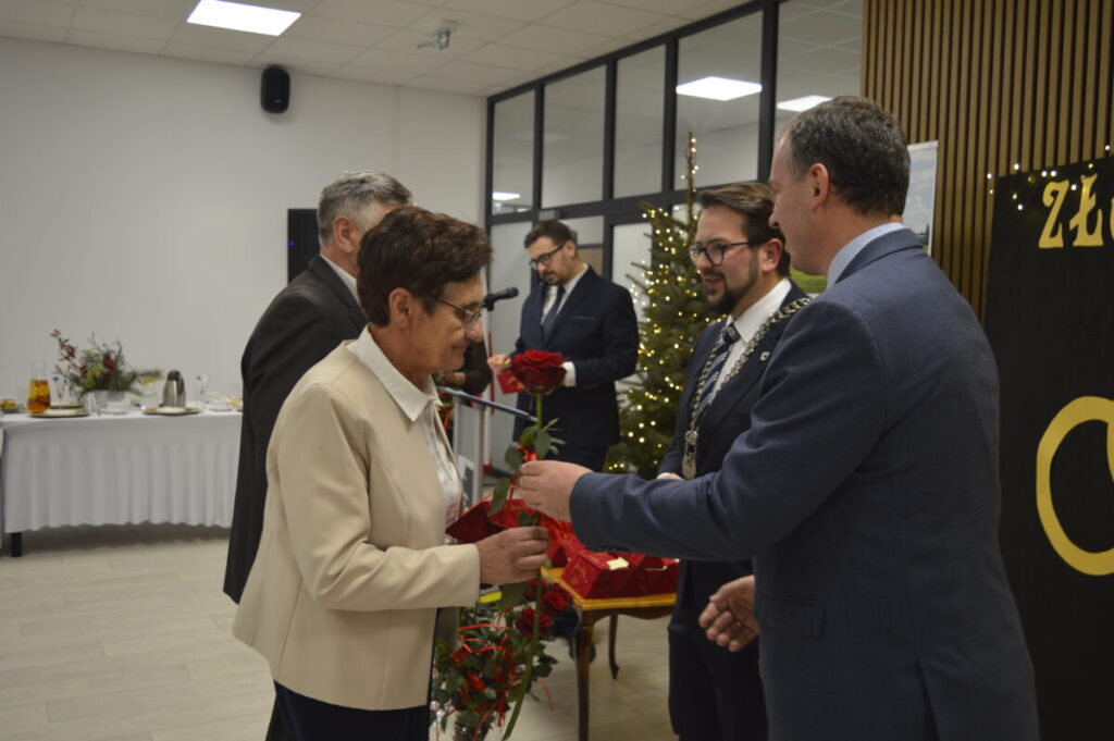 Grupa ludzi w eleganckich strojach wymienia się czerwonymi różami. Tło: choinka ze światełkami i stół z białym obrusem. Przyjęcie w jasnej sali.