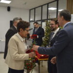 Grupa ludzi w eleganckich strojach wymienia się czerwonymi różami. Tło: choinka ze światełkami i stół z białym obrusem. Przyjęcie w jasnej sali.