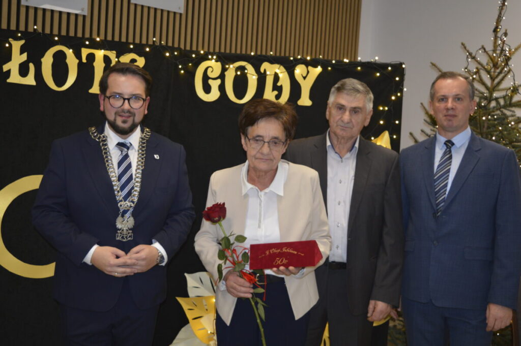 Cztery osoby pozują przed czarnym tłem z napisem Złote Gody. Kobieta trzyma czerwoną różę i pudełko. Obok choinka.