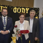 Cztery osoby pozują przed czarnym tłem z napisem Złote Gody. Kobieta trzyma czerwoną różę i pudełko. Obok choinka.