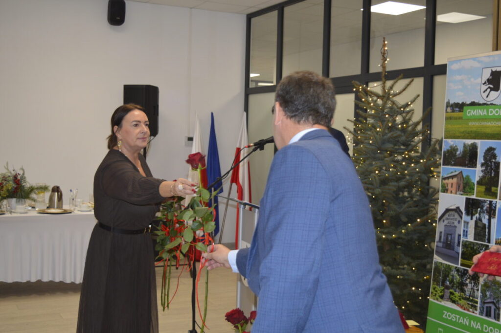 Kobieta wręcza mężczyźnie różę. W tle dekoracje, choinka i baner z napisem Gmina. Dominuje kolor biały i złoty.
