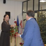 Kobieta wręcza mężczyźnie różę. W tle dekoracje, choinka i baner z napisem Gmina. Dominuje kolor biały i złoty.