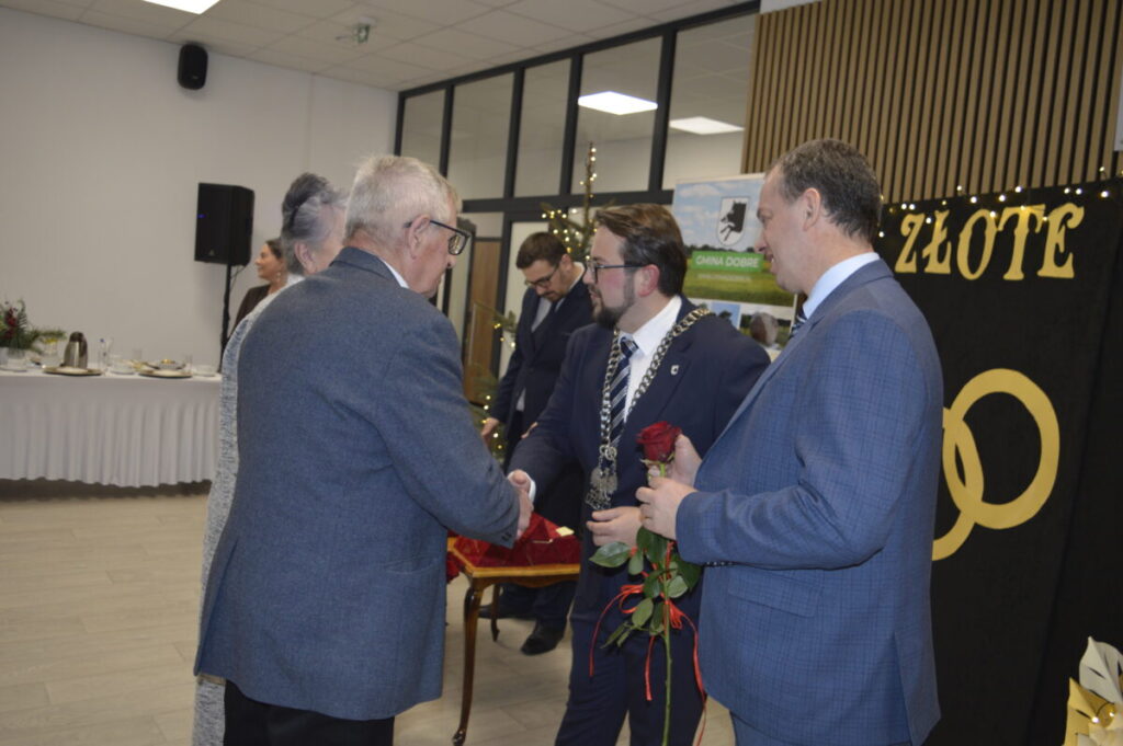 Mężczyzna w garniturze ściska rękę starszemu panu w sali bankietowej. W tle stół z białym obrusem i dekoracje. Dominują odcienie szarości i niebieskiego.