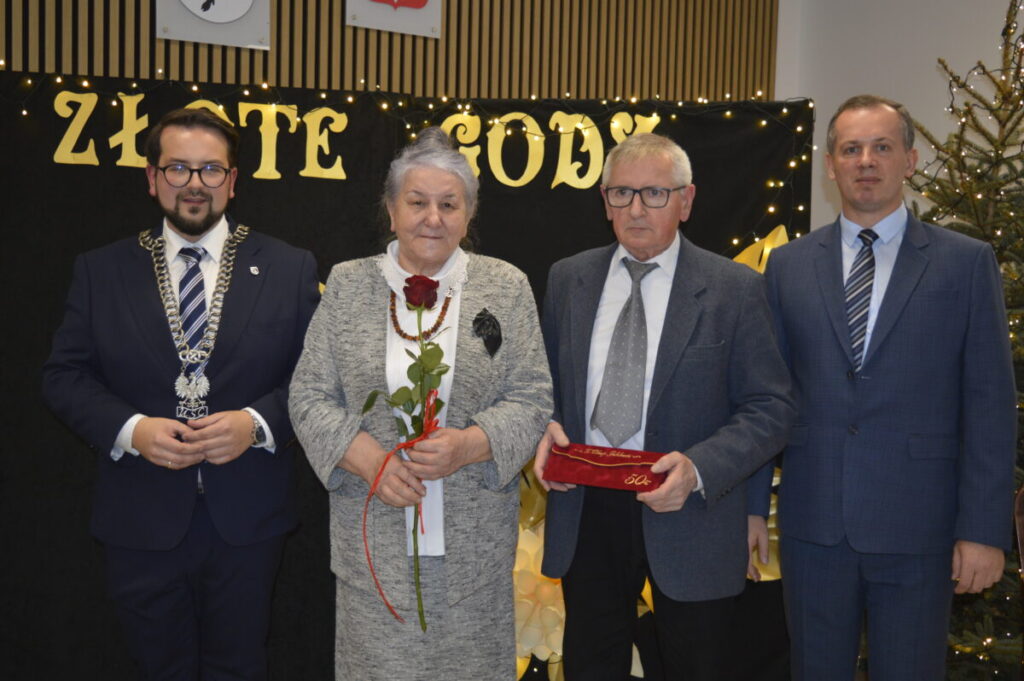 Cztery osoby stoją przed czarnym tłem z napisem ZŁOTE GODY. Starsza kobieta trzyma różę, a obok niej mężczyzna z pudełkiem. W tle choinka i światełka.