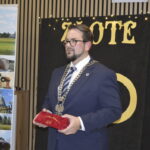 Na zdjęciu widać mężczyznę w garniturze z łańcuchem, trzymającego czerwony aksamitny worek. Tło jest czarne z napisem „ZŁOTE”. Obok stoi banner promujący Gminę Dobre.