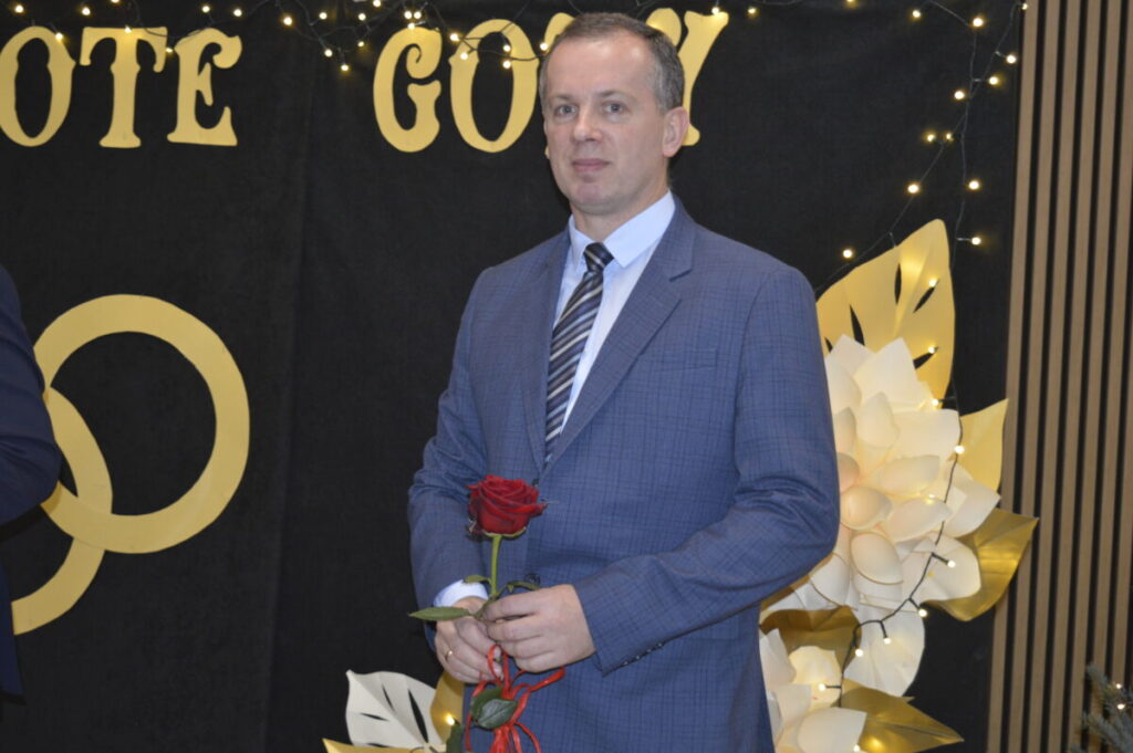 Mężczyzna w garniturze trzyma czerwoną różę. Tło czarne z napisem i złotymi dekoracjami. Obok jasne, papierowe kwiaty.