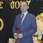 Mężczyzna w garniturze trzyma czerwoną różę. Tło czarne z napisem i złotymi dekoracjami. Obok jasne, papierowe kwiaty.