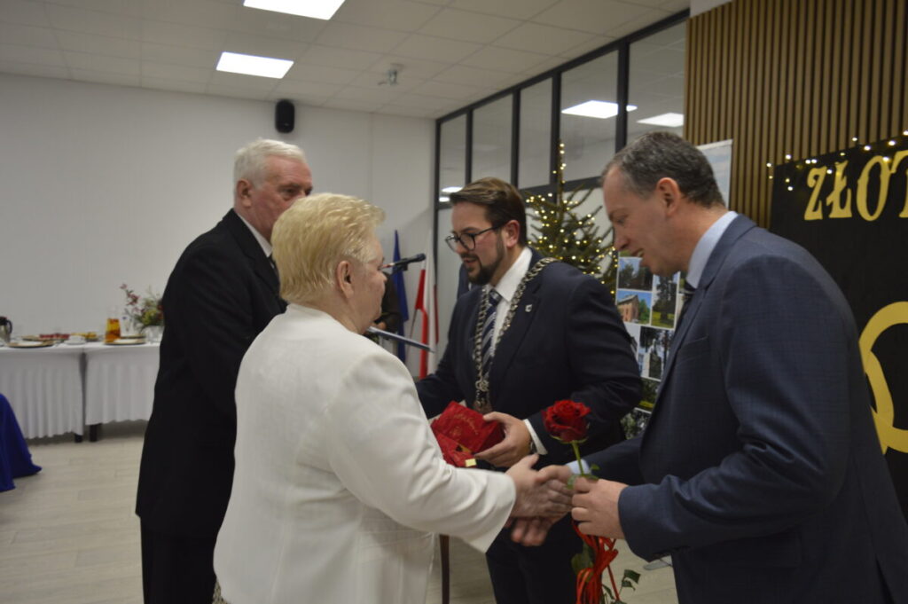 Na obrazie widać osobę starszą w białym ubraniu, która rozmawia z mężczyzną w garniturze. W tle znajduje się choinka z lampkami i stół z przekąskami. Całość ma formalny charakter, z delikatnym oświetleniem.