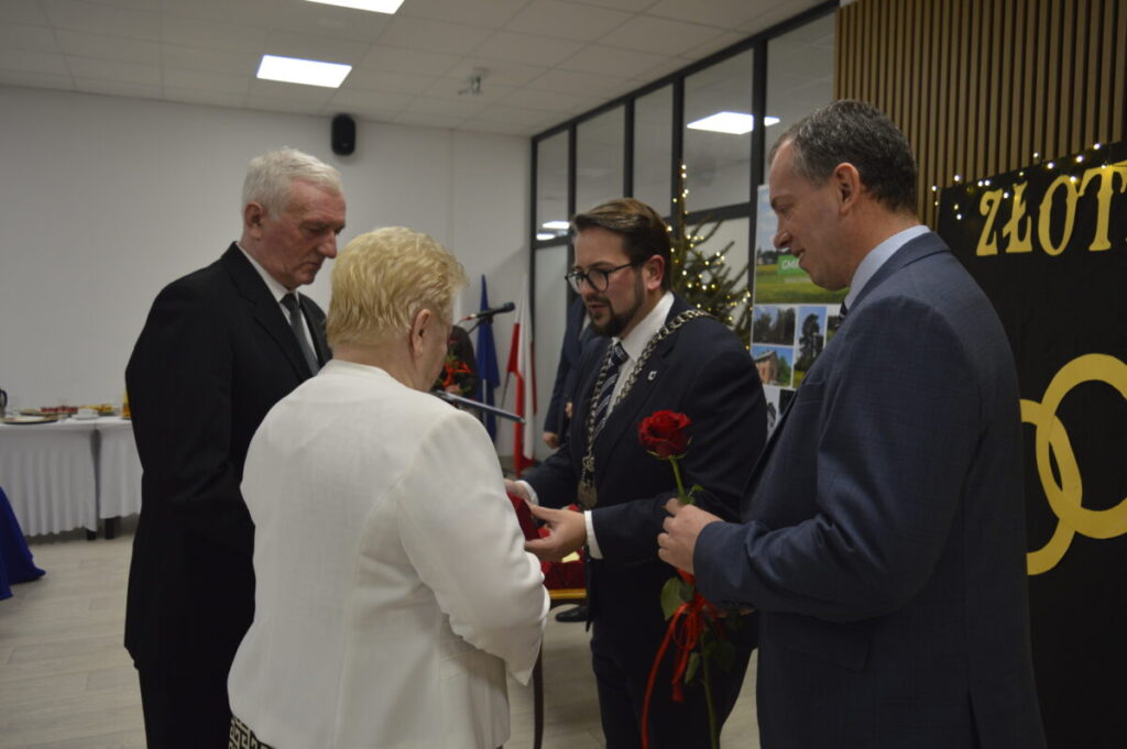 Grupa ludzi podczas uroczystości; mężczyzna z łańcuchem wręcza róże starszej parze. Tło zdobi choinka i złote napisy. Kolory dominujące to czarny, biały, czerwony.