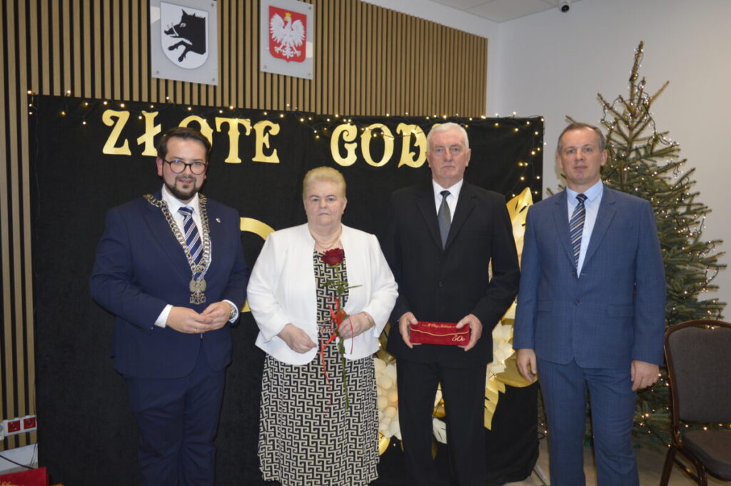 Na zdjęciu są cztery osoby w formalnych ubraniach. Tło jest czarne z napisami ZŁOTE GOD w złotym kolorze. Obok stoi ozdobiona choinka.