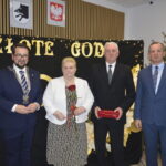 Na zdjęciu są cztery osoby w formalnych ubraniach. Tło jest czarne z napisami ZŁOTE GOD w złotym kolorze. Obok stoi ozdobiona choinka.