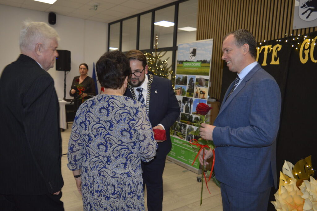 Grupa osób w oficjalnych strojach wymienia się różami, tło dekorowane napisami i choinką. Dominują kolory niebieski i czarny. Atmosfera jest formalna i uroczysta.