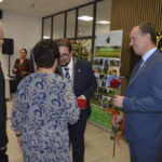 Grupa osób w oficjalnych strojach wymienia się różami, tło dekorowane napisami i choinką. Dominują kolory niebieski i czarny. Atmosfera jest formalna i uroczysta.