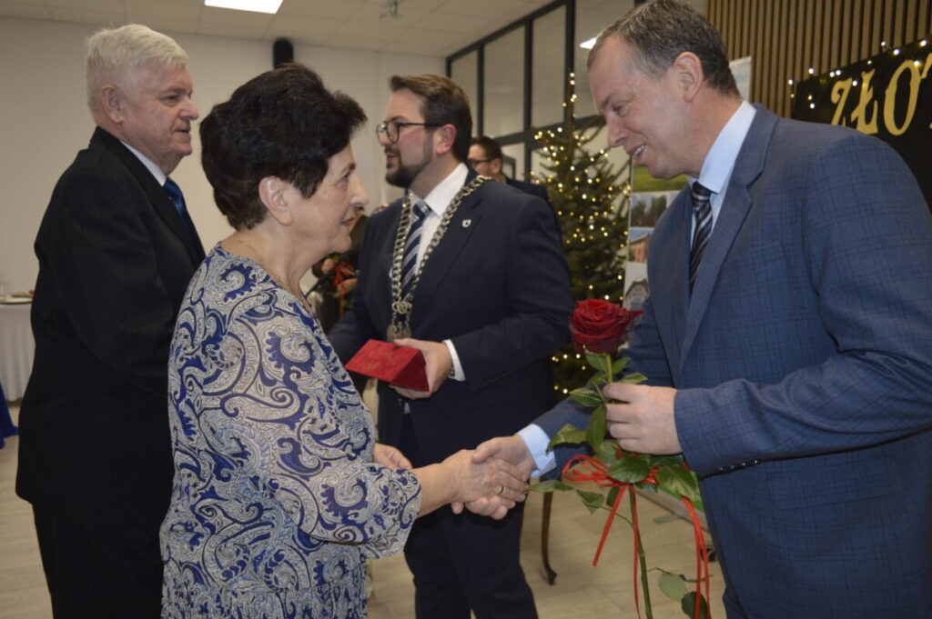 Osoby w eleganckich strojach składają sobie życzenia. Kobieta w niebieskiej bluzce i mężczyzna z czerwoną różą. W tle widać choinkę.