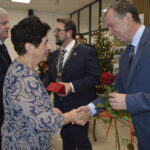 Osoby w eleganckich strojach składają sobie życzenia. Kobieta w niebieskiej bluzce i mężczyzna z czerwoną różą. W tle widać choinkę.