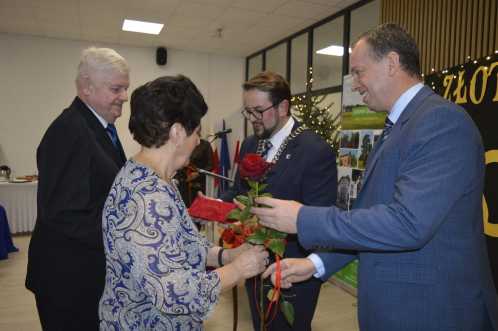 Cztery osoby w formalnym stroju podczas uroczystości. Kobieta z różą, mężczyzna z łańcuchem. W tle dekoracje i tablice.