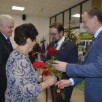 Cztery osoby w formalnym stroju podczas uroczystości. Kobieta z różą, mężczyzna z łańcuchem. W tle dekoracje i tablice.