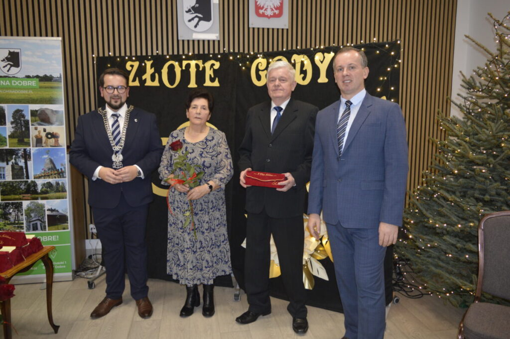 Cztery osoby w eleganckiej odzieży stoją przed czarną ścianką z napisem Złote Gody. Kobieta trzyma bukiet róż, a mężczyzna pudełko. W tle znajduje się choinka z lampkami i baner z krajobrazami.