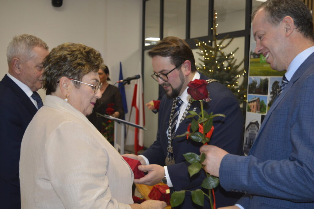 Na zdjęciu znajduje się mężczyzna wręczający kobiecie czerwoną różę. Tło stanowi choinka z lampkami i flagi w biurze. Atmosfera formalna, eleganckie ubrania.