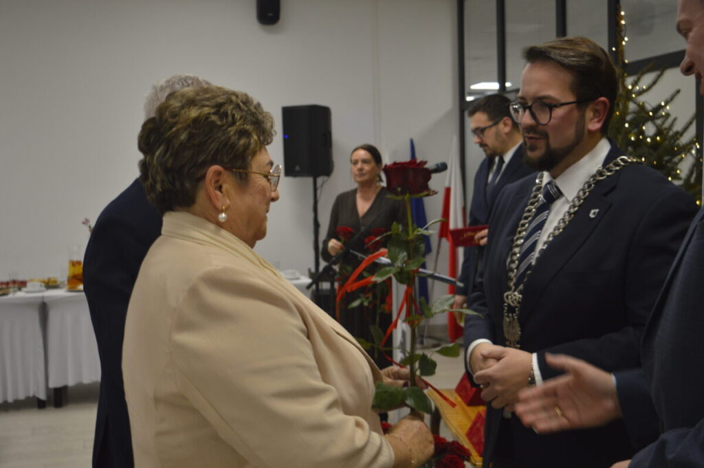 Spotkanie formalne, kobieta w beżowym stroju wręcza różę mężczyźnie w garniturze. Tło jasne, widoczny stół i choinka.