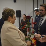 Spotkanie formalne, kobieta w beżowym stroju wręcza różę mężczyźnie w garniturze. Tło jasne, widoczny stół i choinka.