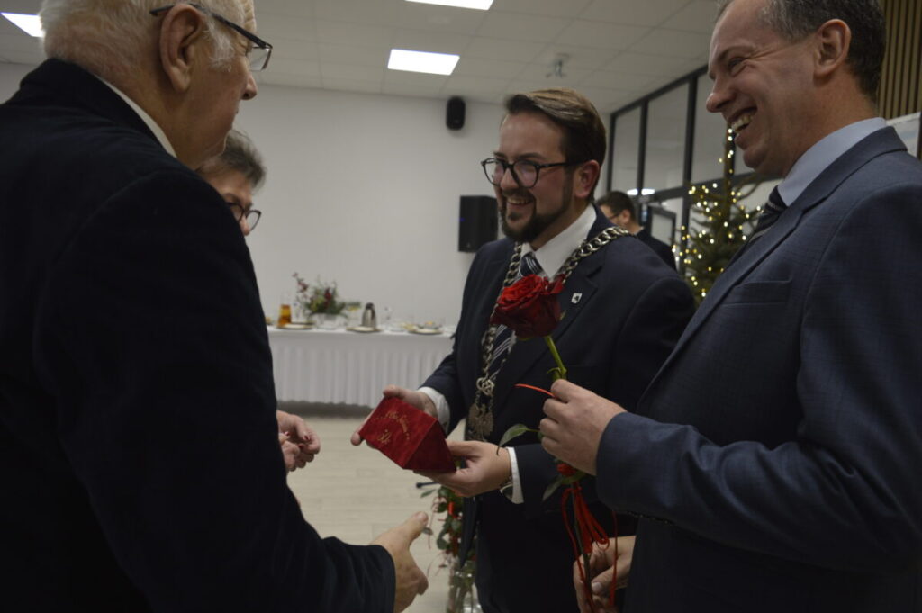 Grupa elegancko ubranych osób w garniturach rozmawia, trzymając róże. W tle jasne pomieszczenie, stół z obrusami i choinka. Atmosfera jest świąteczna i uroczysta.