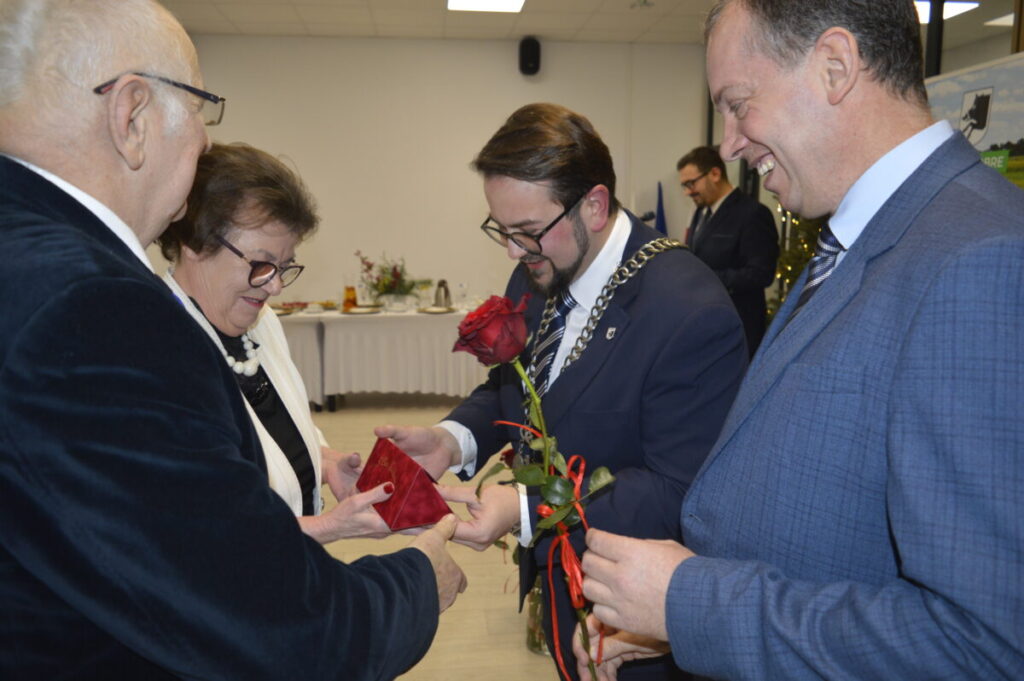 Na zdjęciu grupa osób wymieniających się czerwonym kwiatem i pudełkiem. Tło jasne, z białym stołem i dekoracjami. Ubrani formalnie w garnitury i eleganckie stroje.