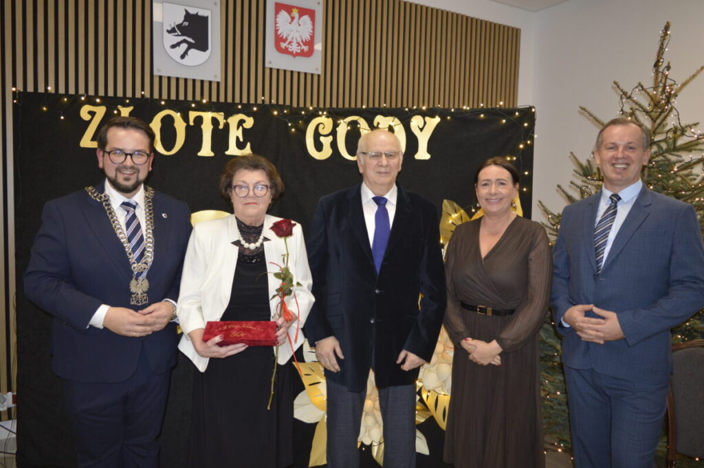 Grupa pięciu osób w eleganckich strojach na tle napisu Złote Gody. Kobieta trzyma różę i czerwoną szkatułkę. W tle choinka i ozdoby.