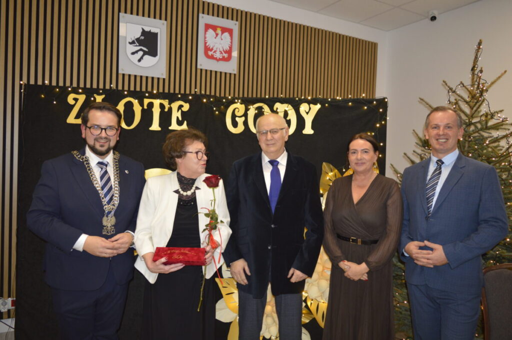 Pięć osób stoi przed czarnym tłem z napisami Złote Gody. Osoby są elegancko ubrane, jedna trzyma czerwoną różę i pudełko. W tle znajduje się choinka oraz godła.