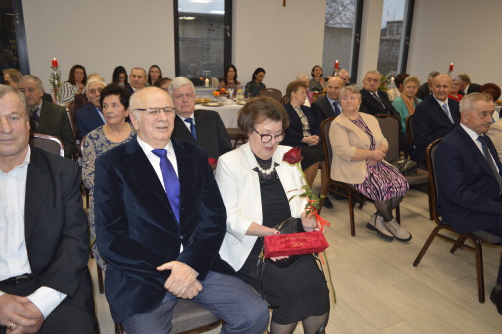 Na zdjęciu grupa starszych ludzi siedzi na oficjalnym wydarzeniu. Pani w białej marynarce trzyma różę. W tle widać świece na stole.