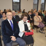 Na zdjęciu grupa starszych ludzi siedzi na oficjalnym wydarzeniu. Pani w białej marynarce trzyma różę. W tle widać świece na stole.