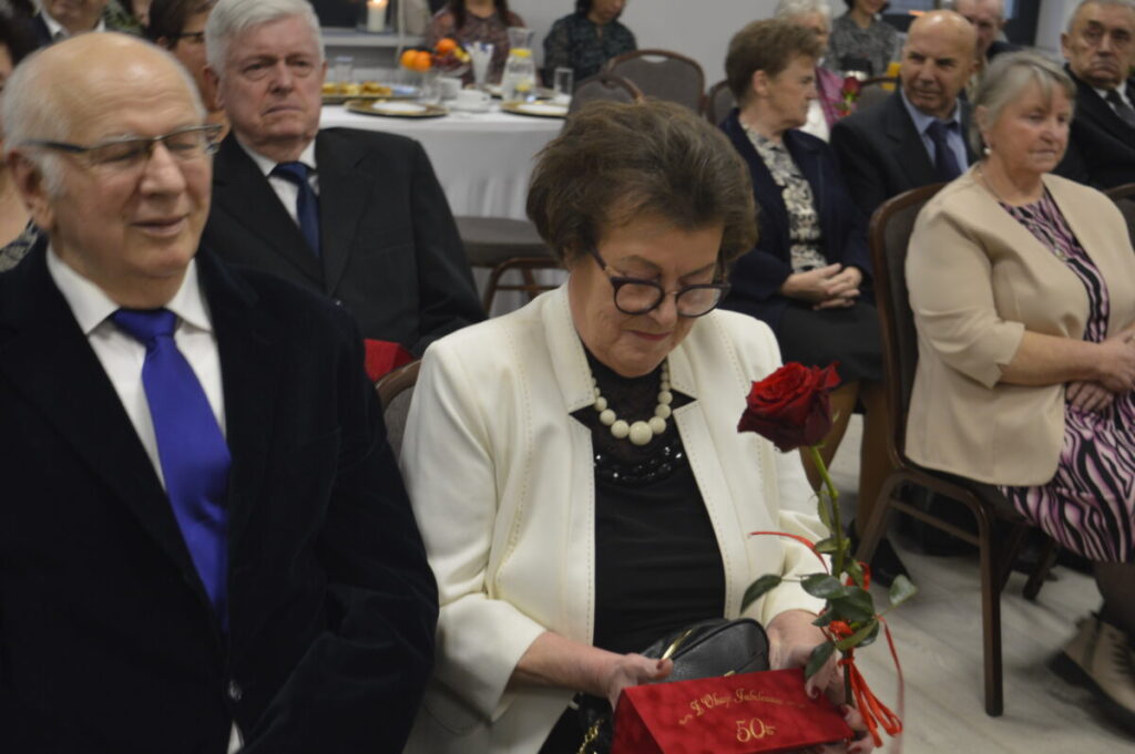 Na zdjęciu widać starszych ludzi siedzących przy stole, kobieta trzyma czerwoną różę. Ubrania są eleganckie, a tło to sala bankietowa. W oddali widać nakryte stoły.