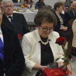 Na zdjęciu widać starszych ludzi siedzących przy stole, kobieta trzyma czerwoną różę. Ubrania są eleganckie, a tło to sala bankietowa. W oddali widać nakryte stoły.