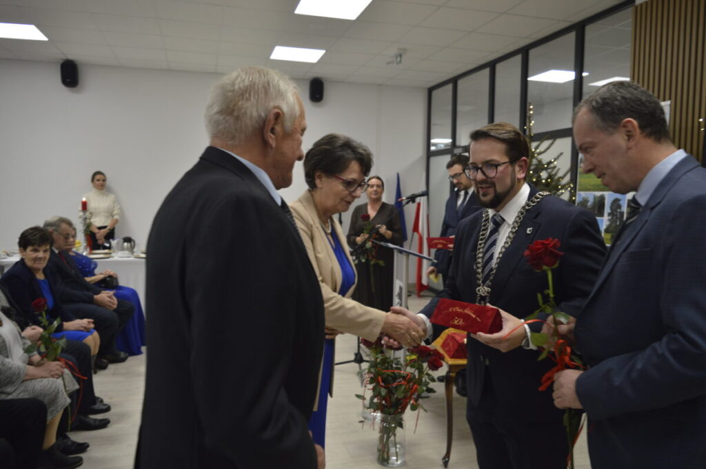 Na zdjęciu kilku elegancko ubranych ludzi, trzymają kwiaty, wręczając nagrody w jasno oświetlonym pomieszczeniu. Obok choinka. Na ścianie flagi.