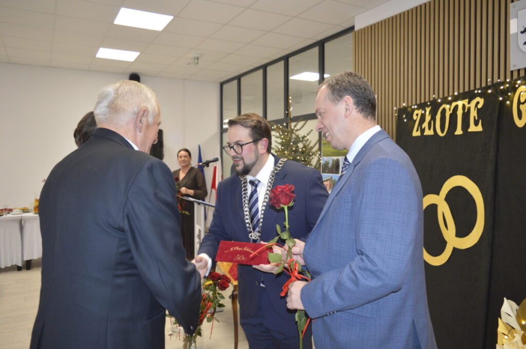 Na zdjęciu widnieje grupa osób w eleganckich strojach podczas uroczystości. Tło jest jasne z dekoracją ZŁOTE GODY i kwiatami. Osoby wymieniają uściski dłoni i wręczają róże.